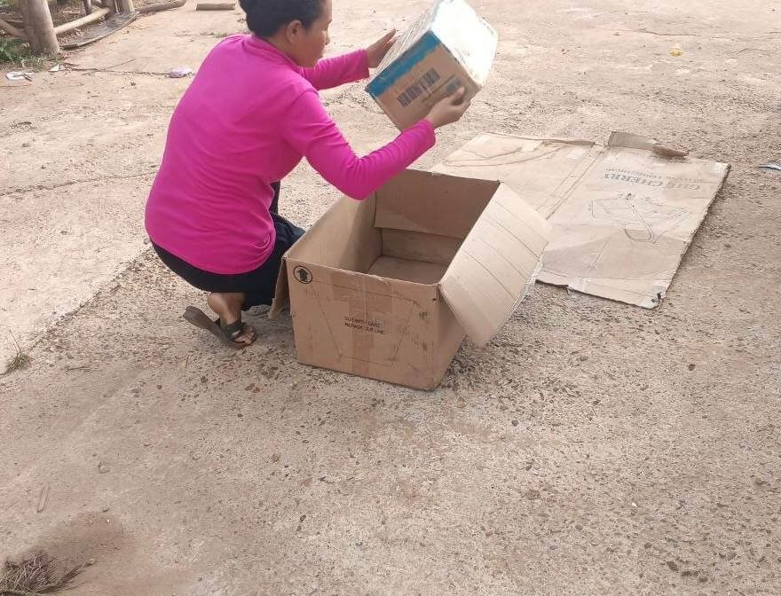A mother placing a small cardboard box inside a larger rectangular box to build a DIY car on a concrete path.