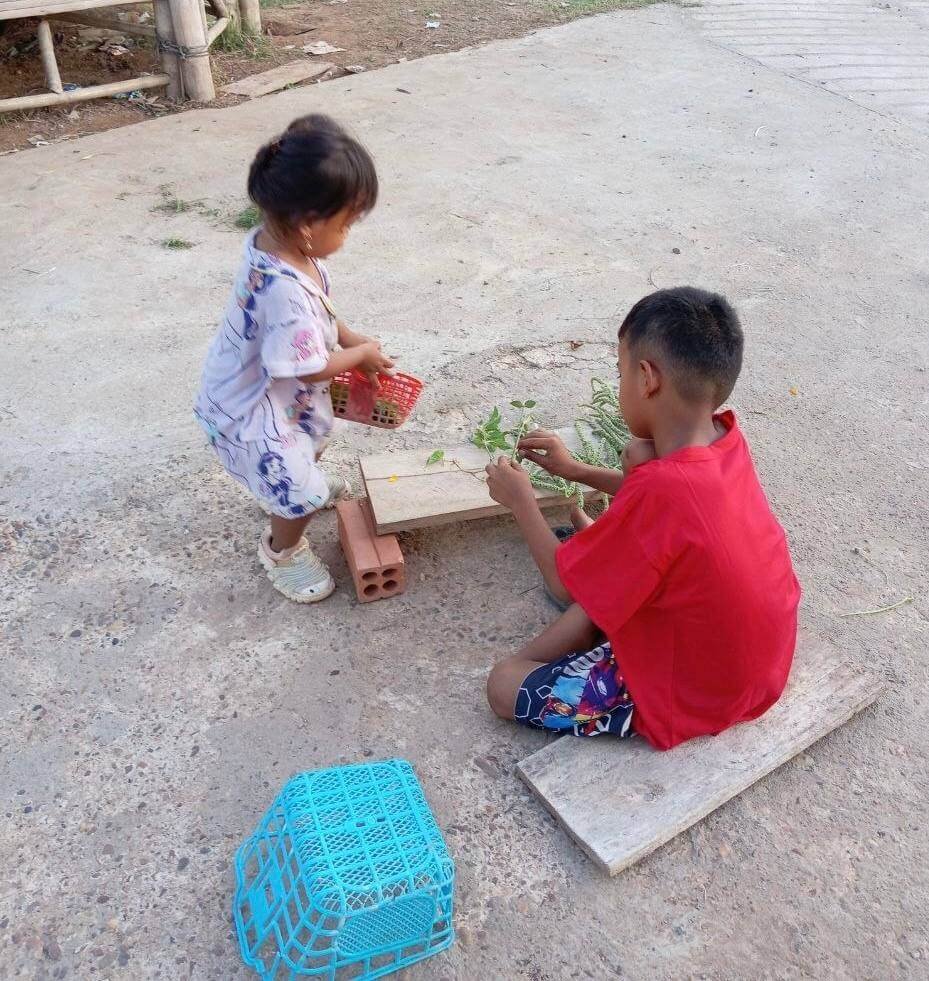 Two siblings setting up a pretend shop using wooden boards, bricks, and plastic baskets.