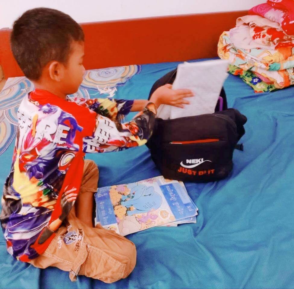 An 8-year-old boy sitting on his bed packing notebooks into his black school bag.