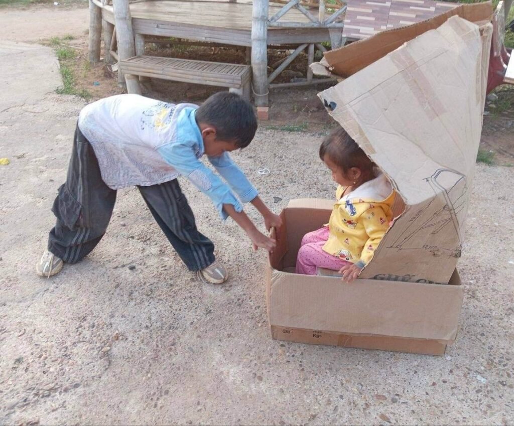 An older brother pushing his younger sister sitting inside a cardboard box car along a concrete garden path.