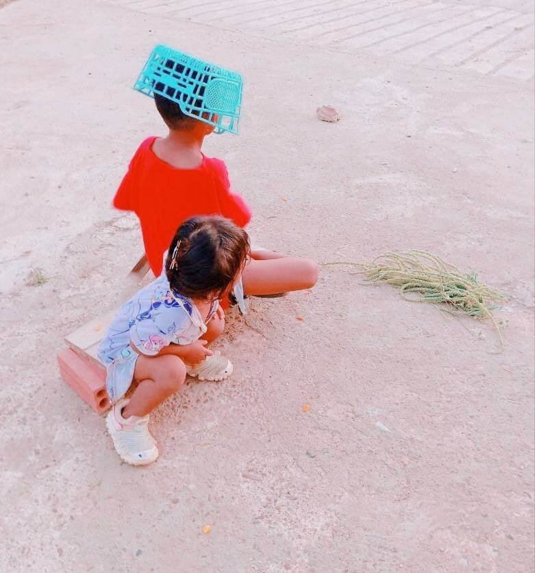 A boy wearing an upside-down blue plastic basket on his head as a makeshift helmet while pretending to ride a bike with his sister.