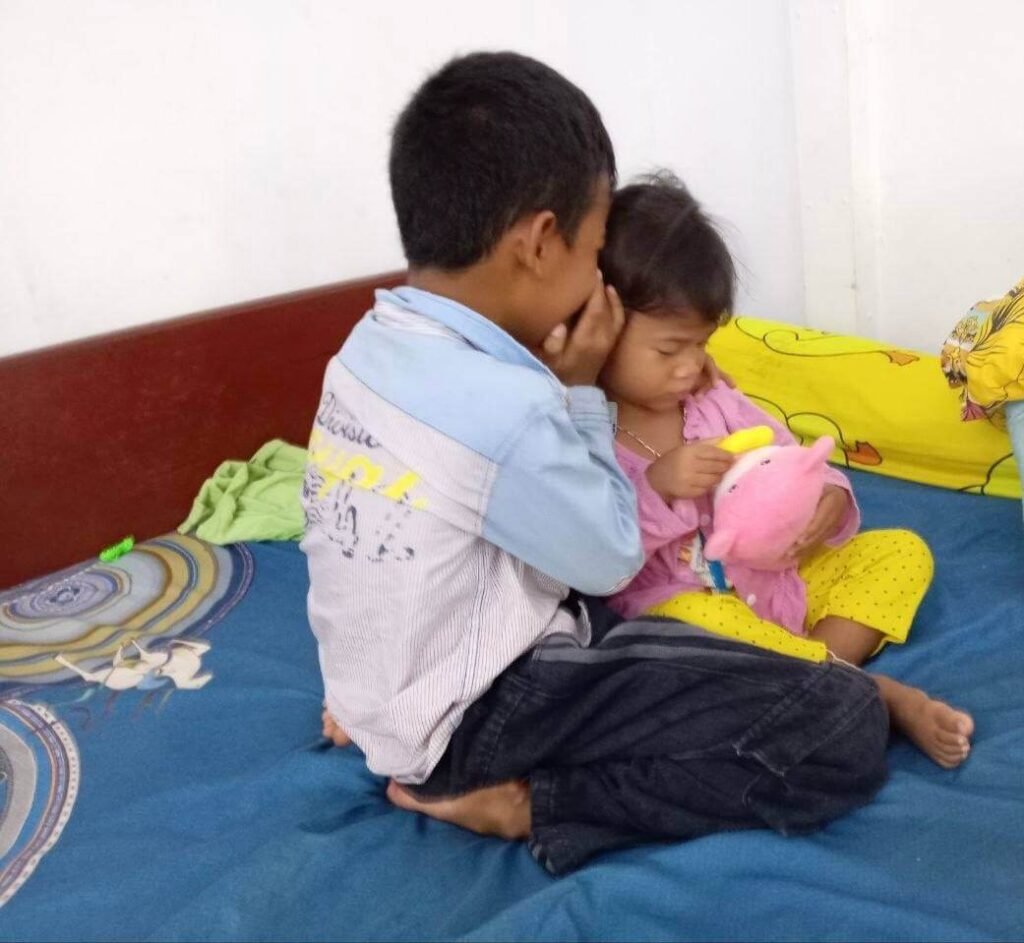 An older brother leans in closely to whisper a secret to his younger sister while they both sit on a bed.