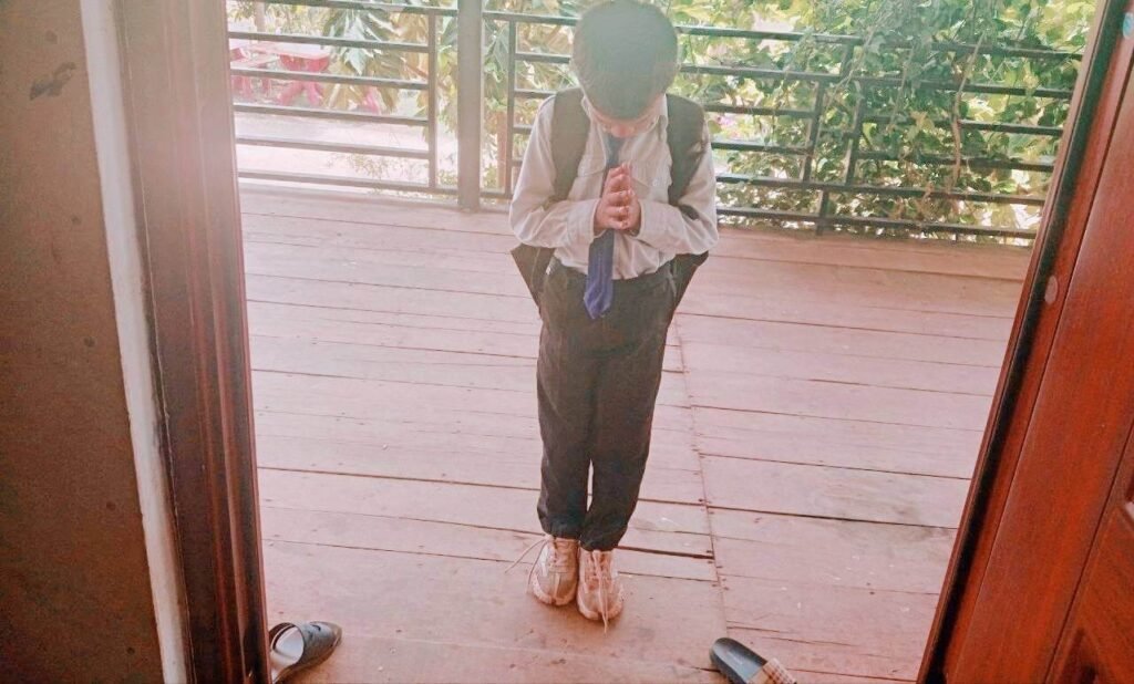 A boy in a school uniform standing by the door performing the traditional Cambodian Sampeah greeting.