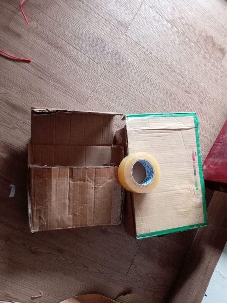 Two empty cardboard boxes and a roll of clear packing tape on a wooden floor.