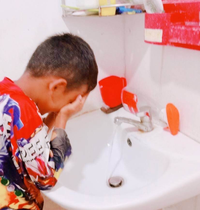 A young boy washing his face at the bathroom sink to wake up.
