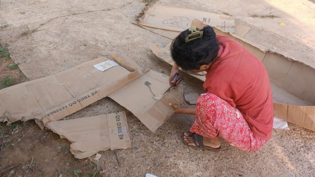 A person kneeling outside cutting a large cardboard box with scissors to create a boat shape.