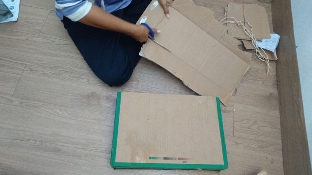 A person sitting on the floor using scissors to cut straps out of a flat piece of cardboard.
