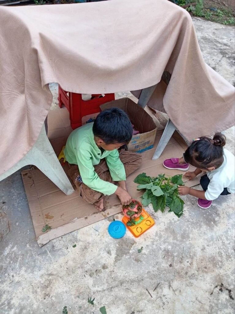 Two children sitting on a cardboard box under a DIY outdoor table fort made with a tan blanket and propped up by a small red stool.