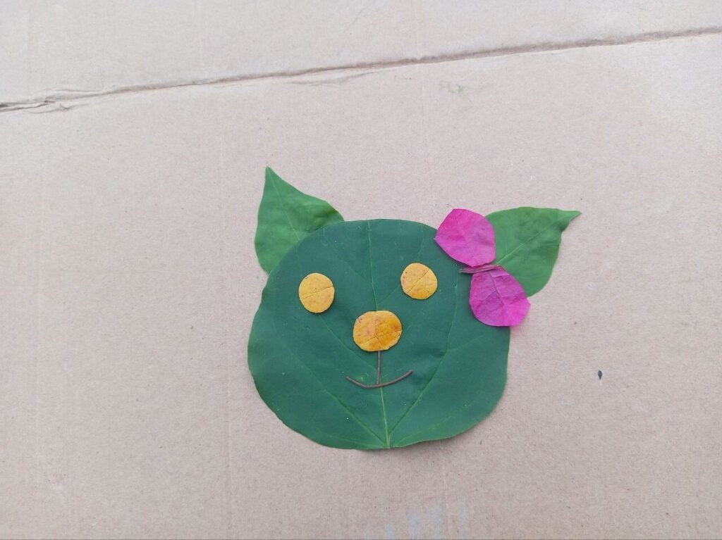 Finished leaf animal faces craft showing a green leaf cat face with yellow petal eyes and pink flower bow.