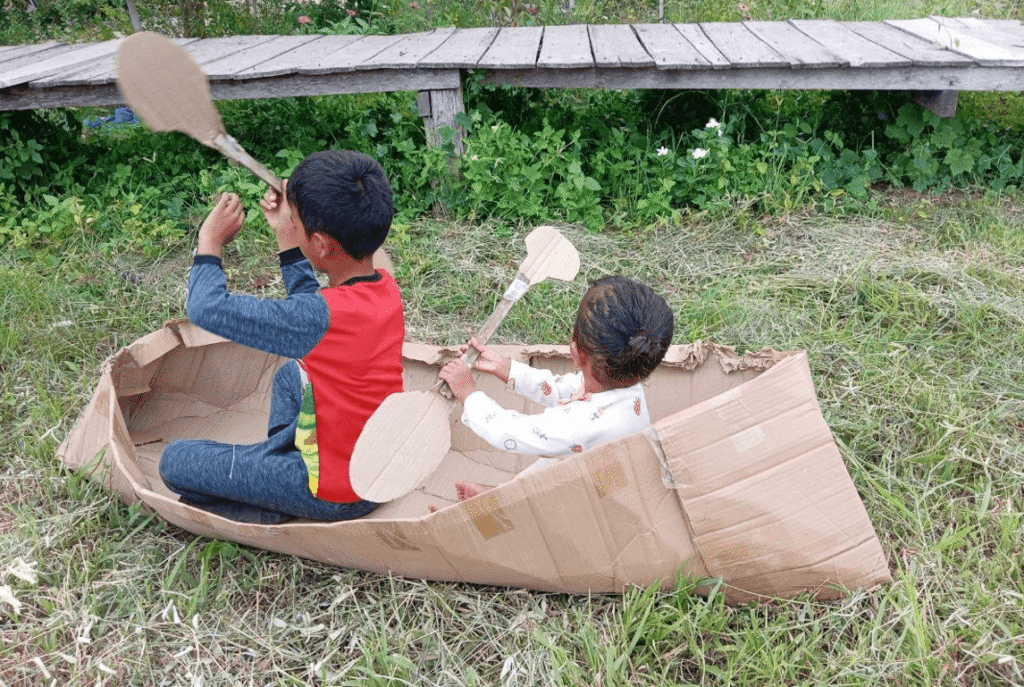 A young boy and toddler girl sitting inside a DIY cardboard boat on the grass, holding cardboard oars.