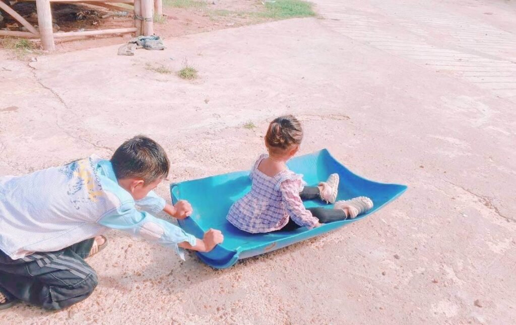 An older brother pushing his toddler sister in a DIY ride-on toy made from a blue plastic barrel.