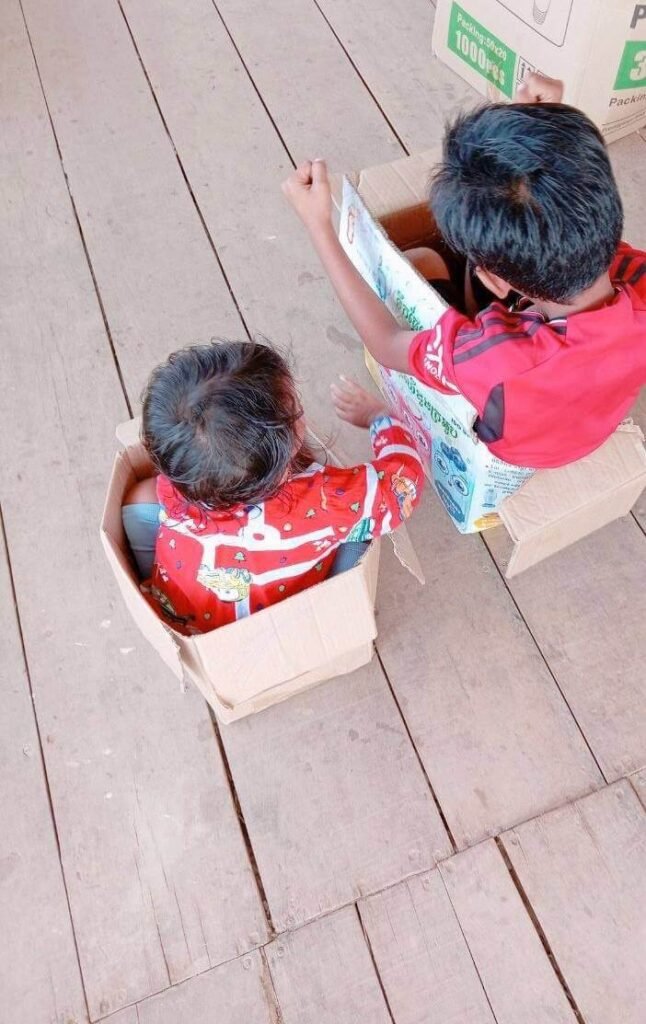 Two children sitting in separate cardboard boxes on a wooden floor getting ready for a pretend race.