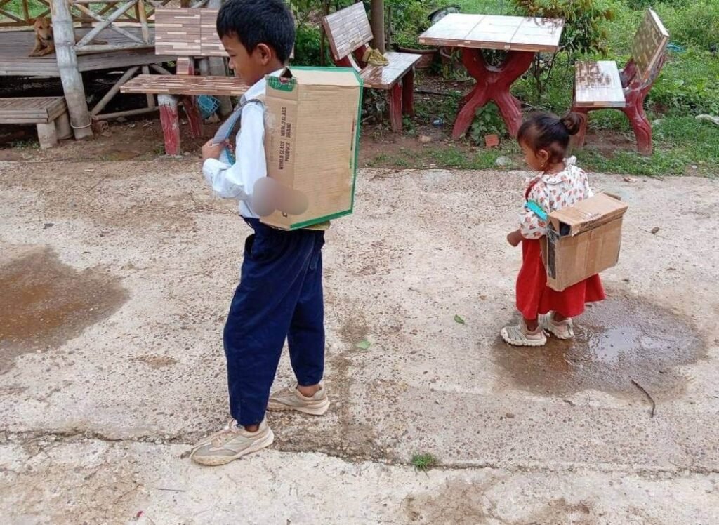 A young boy in uniform and a toddler girl in a red dress wearing homemade cardboard backpacks outside.