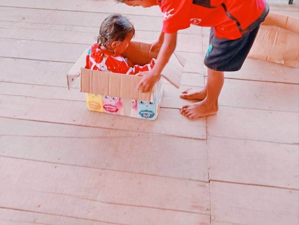 An older brother pulling his toddler sister across a wooden floor while she sits inside a cardboard delivery box.