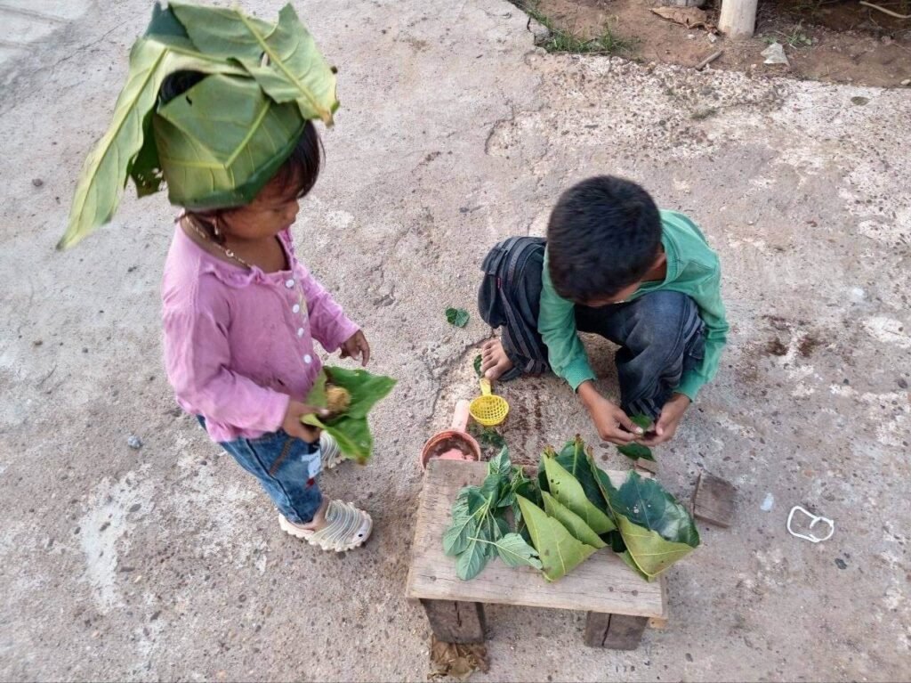Toddler girl wearing a large green leaf as a chef hat while her brother plays with leaves and a strainer.