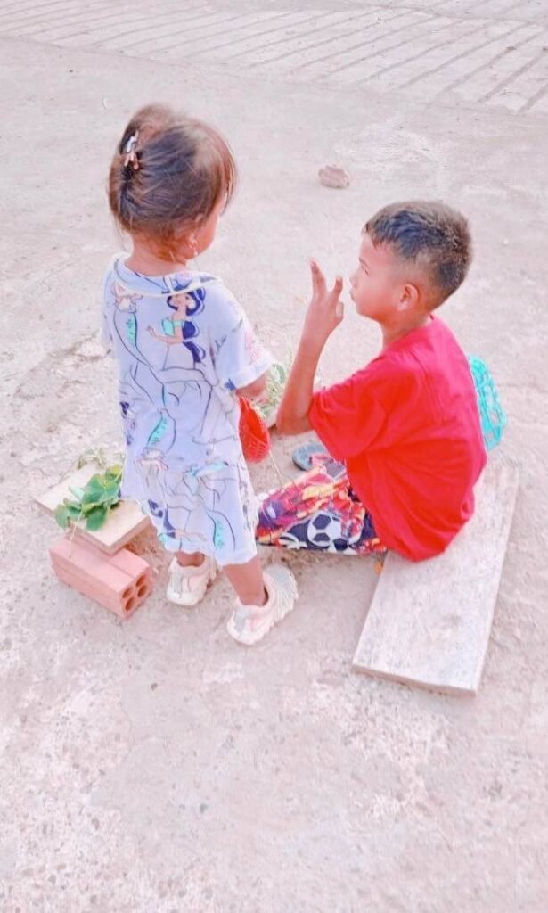 An older brother holding up three fingers to show his toddler sister the price of her vegetables during pretend play.
