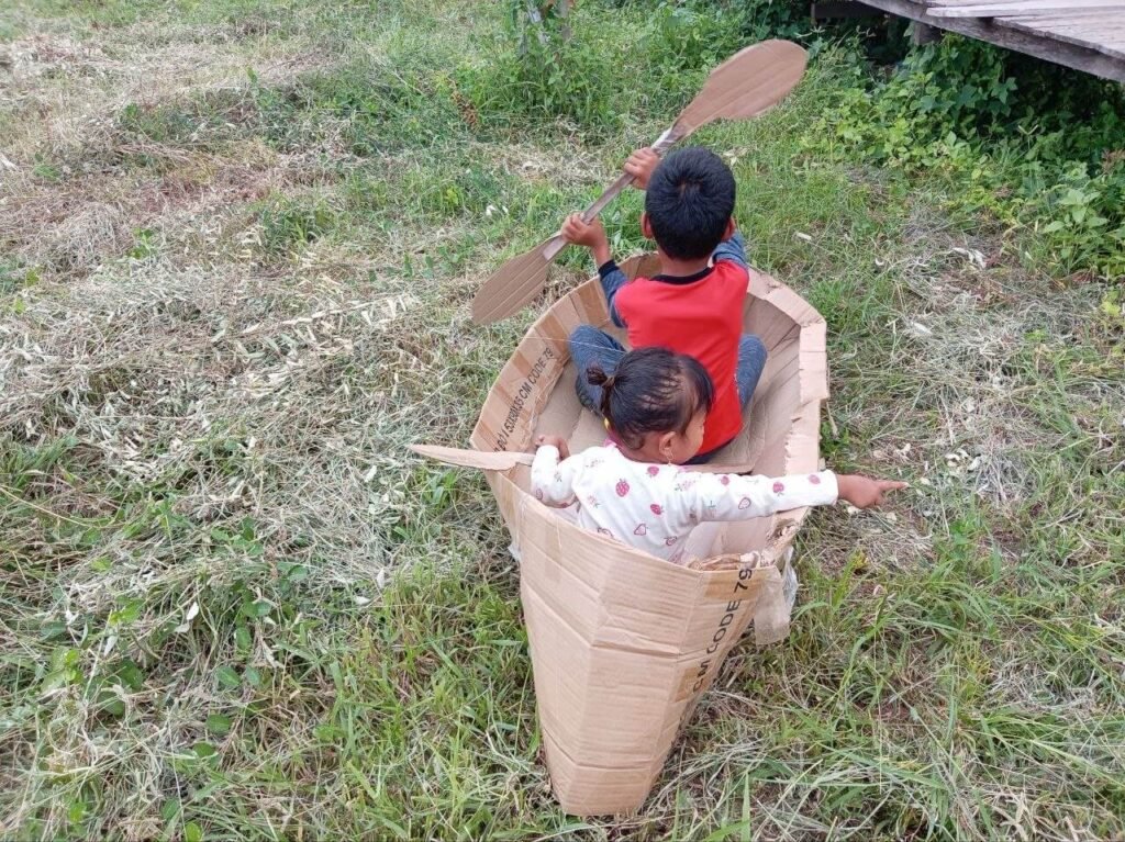Top-down angle of two children rowing a homemade cardboard boat on the lawn.
