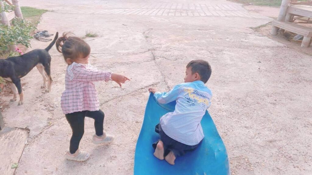 A boy sitting in a blue plastic barrel toy while his younger sister stands and points, asking for her turn back.
