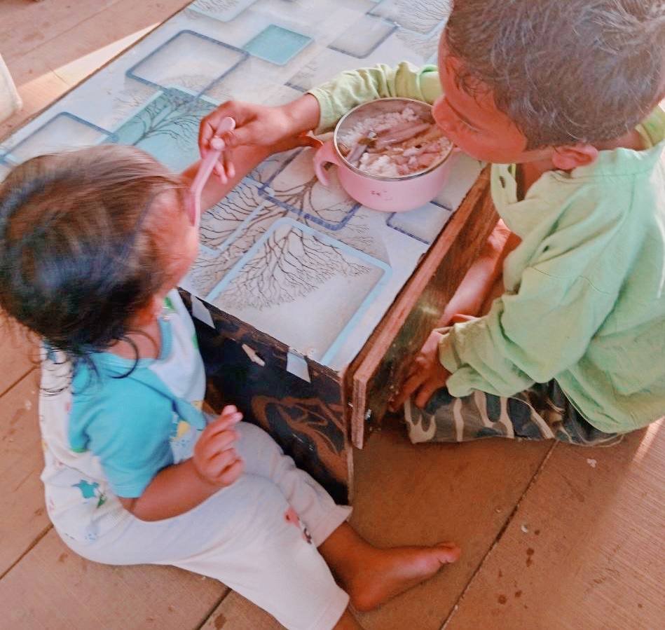 A young boy and his toddler sister sitting together sharing a meal out of a pink bowl.
