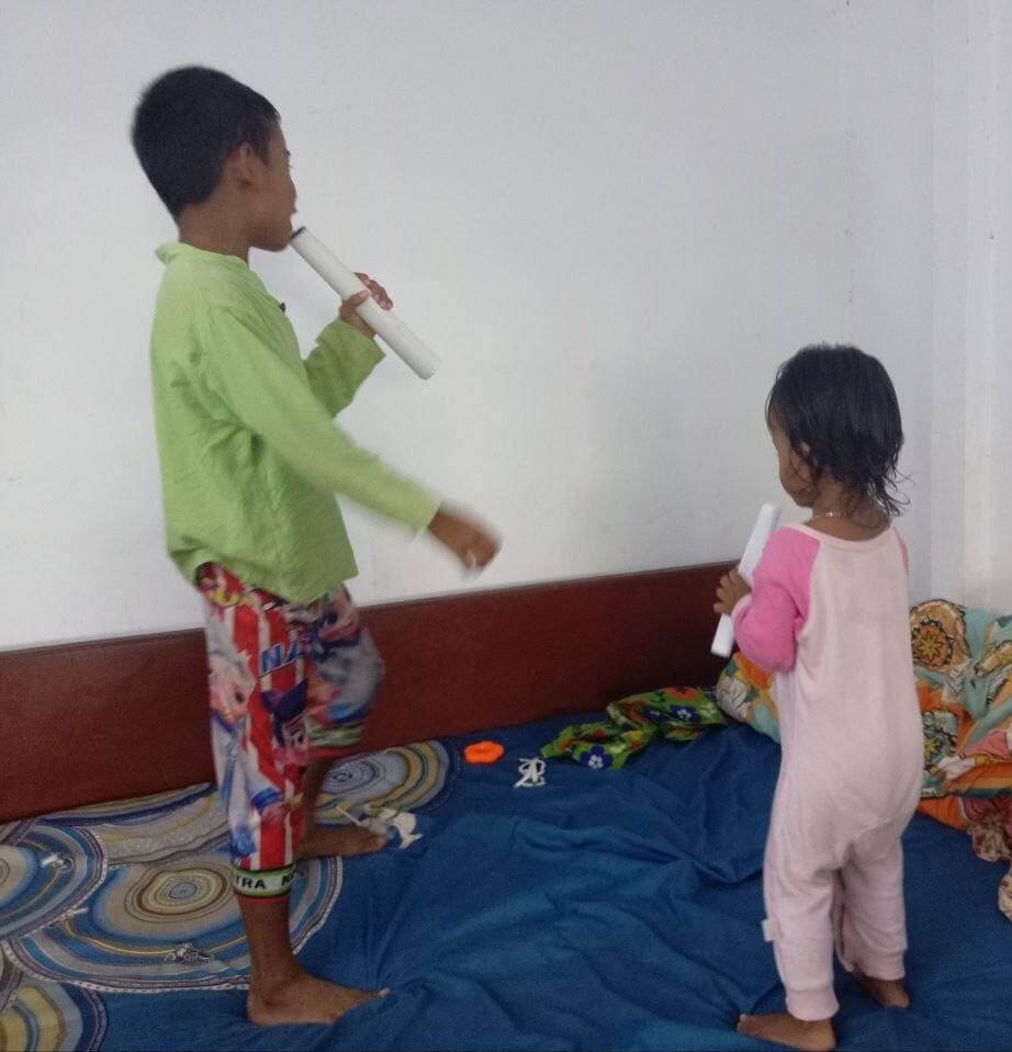 Two children, a boy and a girl, standing on a bed holding paper rolls as microphones and singing together at night.