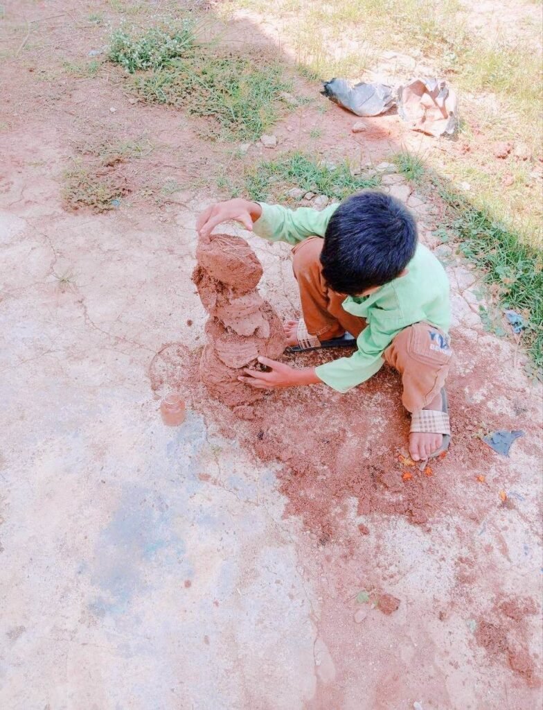 A young boy crouching in the dirt stacking large mud clods into a tall tower.