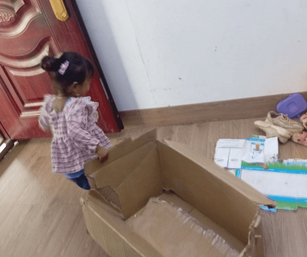 A toddler girl walking toward a large empty cardboard box on a wooden floor.