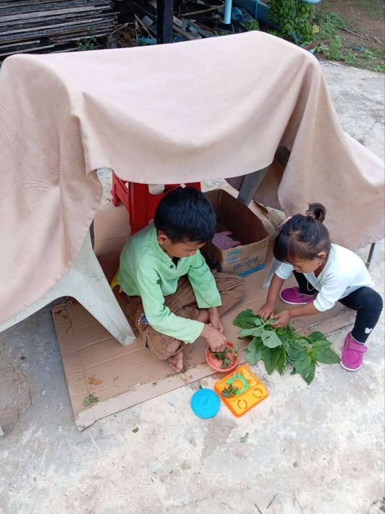 An 8-year-old boy and a 3-year-old girl making pretend soup with green leaves and a plastic toy stove inside a backyard table tent.