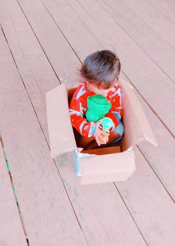 A toddler girl sitting quietly inside a cardboard box playing with green and orange toys.