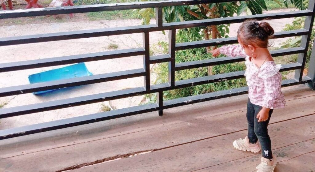 A toddler girl standing on a patio pointing down at a blue half-cut plastic barrel in the yard.