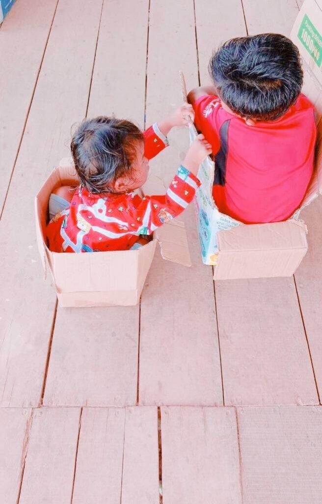 A toddler girl reaching from her cardboard box to touch her older brother's box during pretend play.