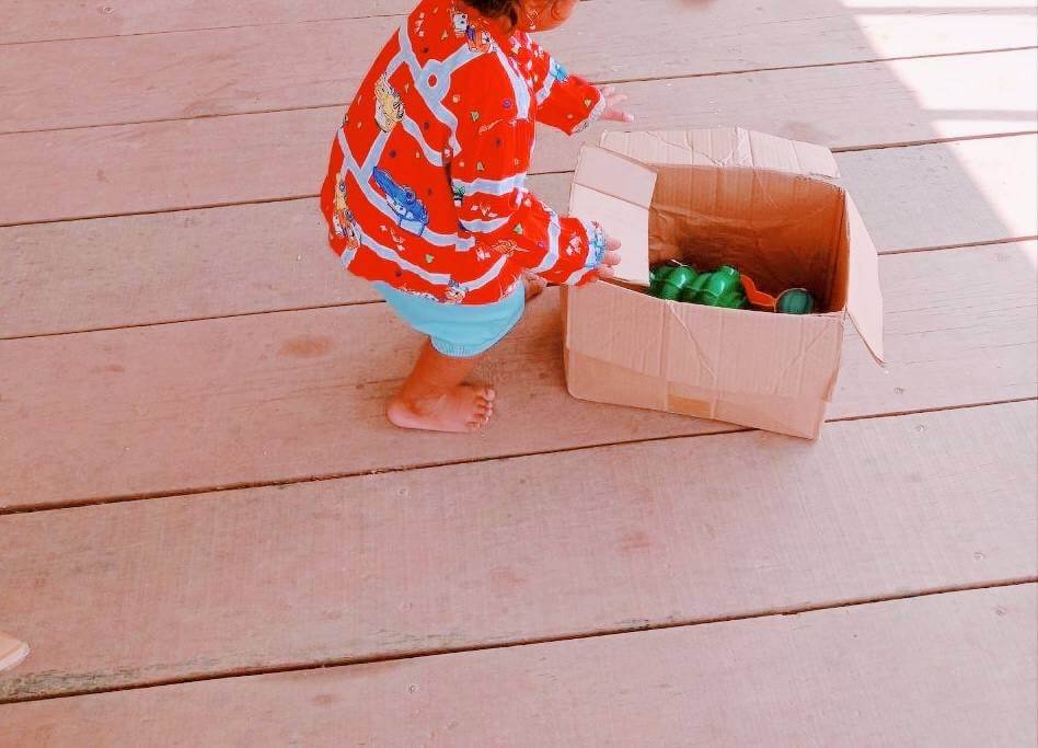A toddler girl standing on a wooden floor placing green plastic toys into an empty cardboard box.