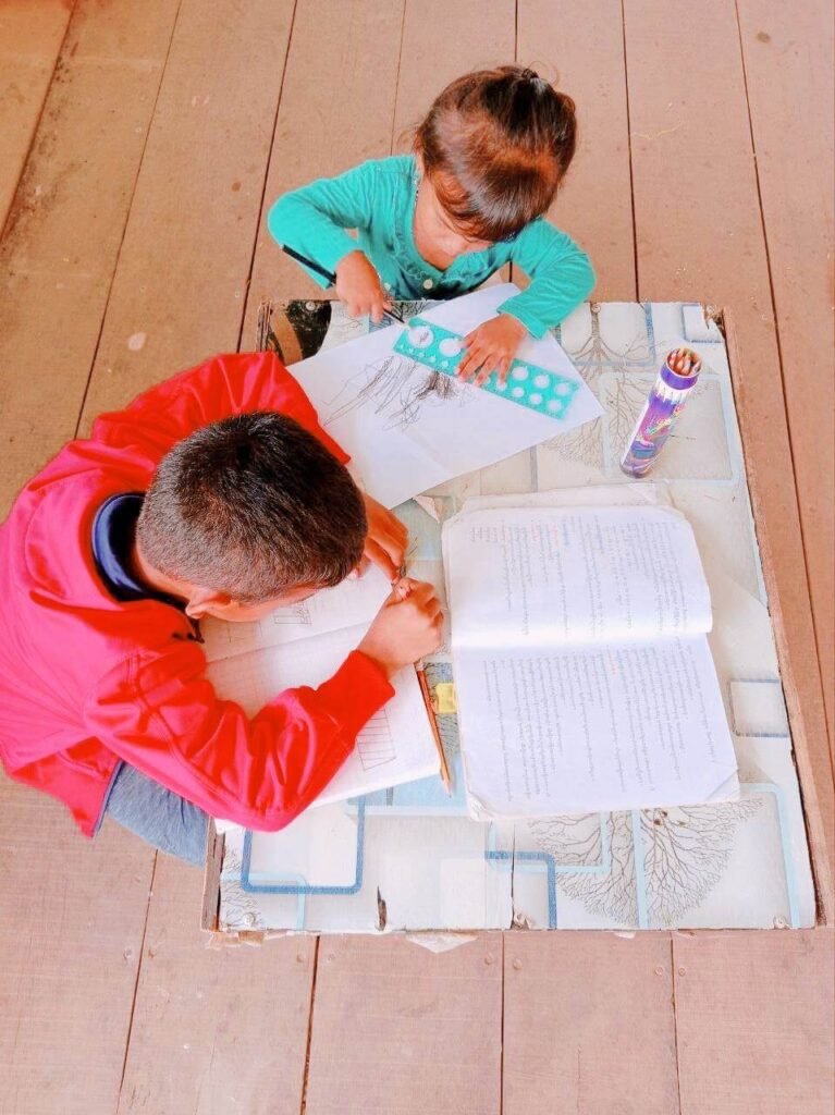 Top-down view of an older boy doing homework while a toddler girl draws next to him at a low table.