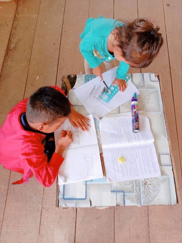A toddler girl pressing a blue plastic stencil ruler onto blank paper while her brother writes.