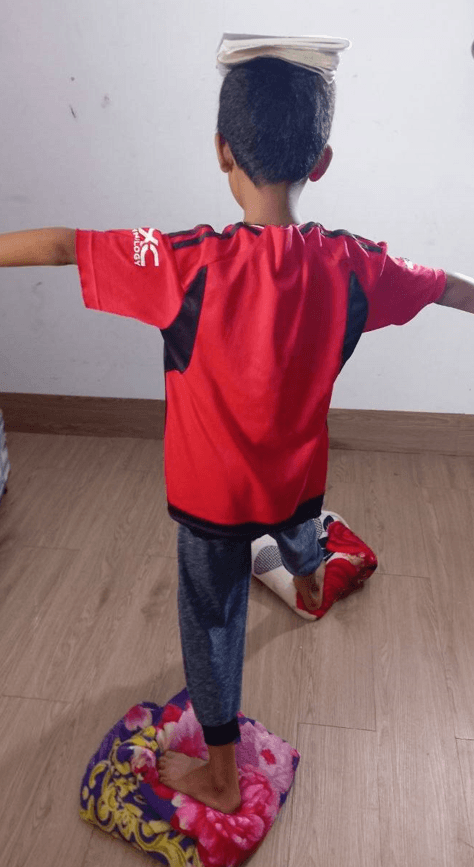 A young boy balancing a book on his head while standing on one foot on a folded blanket.
