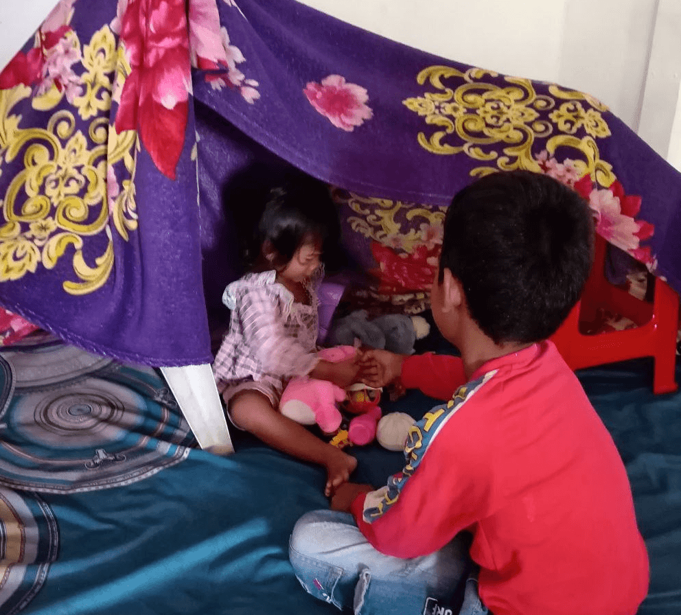 Two children sitting inside a homemade fort made from a purple blanket draped over chairs.