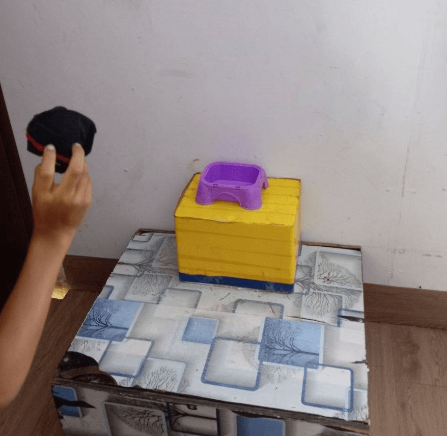A hand throwing a rolled-up black sock into a small purple bowl sitting on a yellow box.